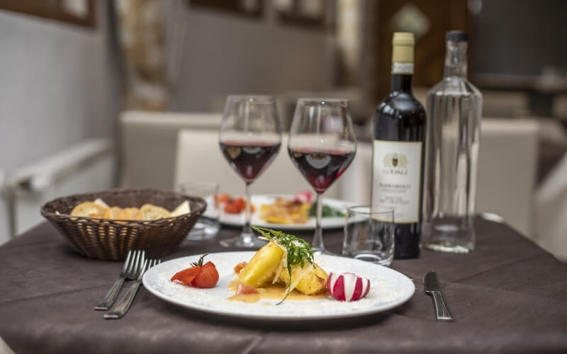 Table set for dining with wine glasses, a yellow vegetable dish, cherry tomatoes, radish, and a wicker basket of bread.