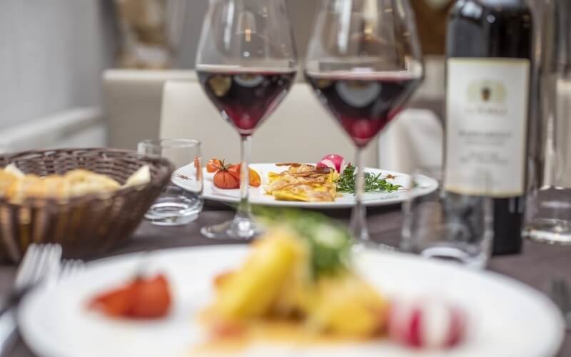 Close-up of a table set with pasta, vegetables, bread, wine glasses, and a wine bottle in a cozy restaurant setting.