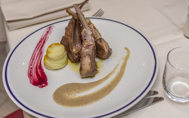 Plate of four perfectly cooked lamb chops with sliced potatoes, red and brown sauces, elegant table setting in background.