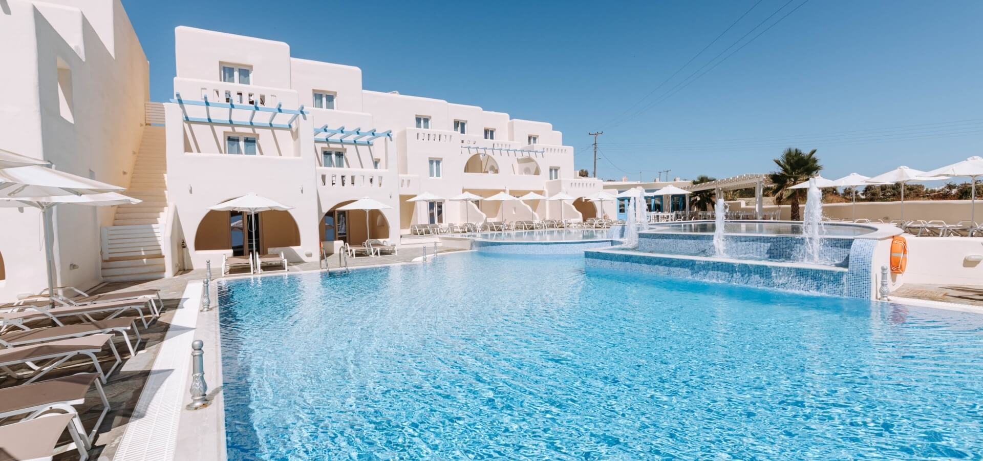 Una piscina serena con fontana, lettini e ombrelloni, circondata da un edificio bianco sotto un cielo blu.