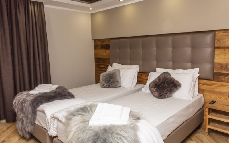 Hotel room with two beds, gray leather headboard, wooden nightstand, recessed lighting, and light hardwood floor.