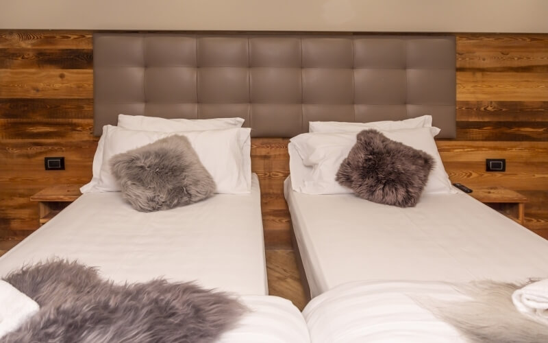 Cozy bedroom with two side-by-side beds, white bedding, gray fur blankets, and rustic wooden walls, creating a serene atmosphere.