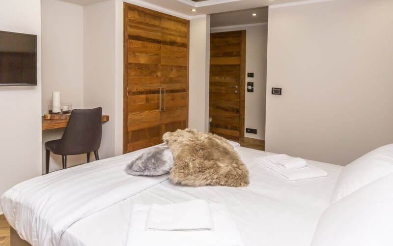 Serene bedroom with neutral tones, a central bed, wooden desk, TV, and warm lighting, creating a cozy atmosphere.