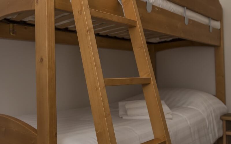 Wooden bunk bed with white bedding, ladder on the left, and a small stool on the right against a plain white wall.