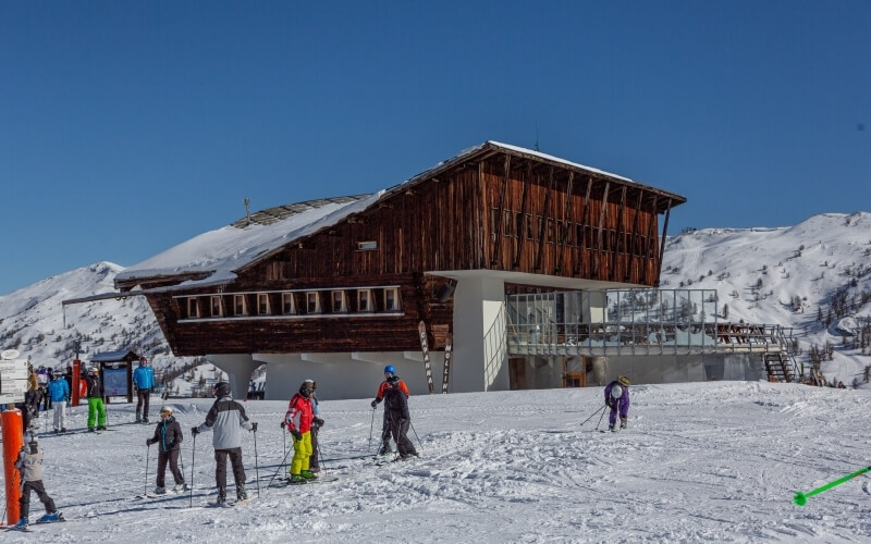 A group of people in winter attire prepares to ski on a snowy slope in front of a large wooden and stone building.