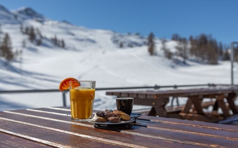 A wooden table outdoors holds a glass of orange juice, a black cup, and cookies, with snow-covered mountains in the background.