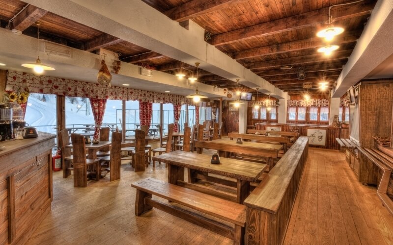 Rustic wooden dining area with tables, benches, a long counter, hanging lights, and large windows showing a snowy landscape.