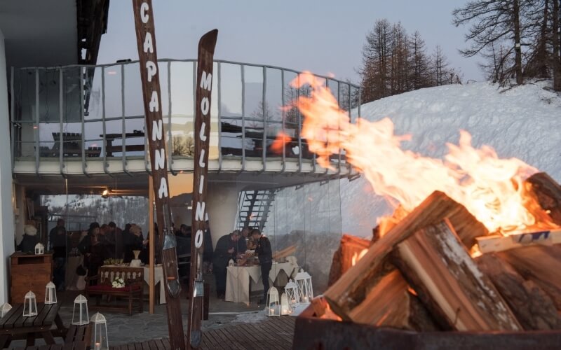 Winter scene with a bonfire, skis, lanterns, a restaurant, and a mountain slope under a clear blue to grey sky.