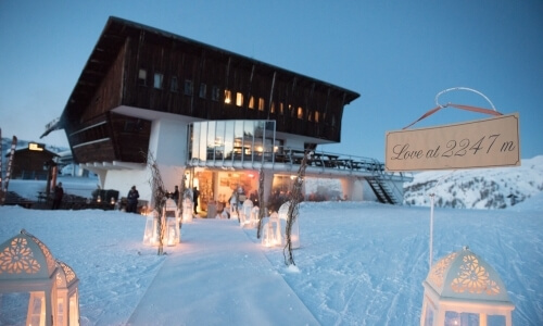 Snowy scene featuring a wooden building, a "Love at 2247m" sign, and a group of warmly dressed people outside.