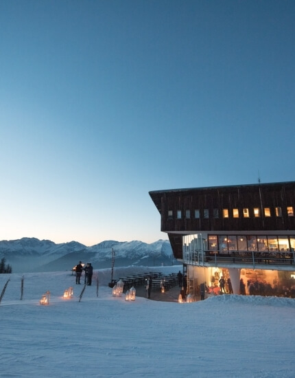 Modern building on snowy hill with large windows, balcony, and people outside, set against a stunning mountain backdrop.