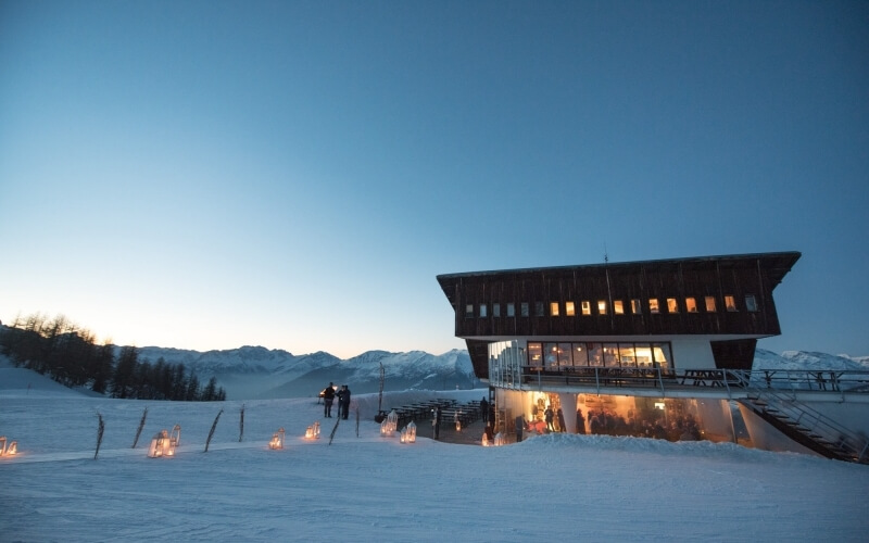 Serene winter scene of a two-story building with a large glass window, surrounded by snow and mountains, warm light glowing inside.