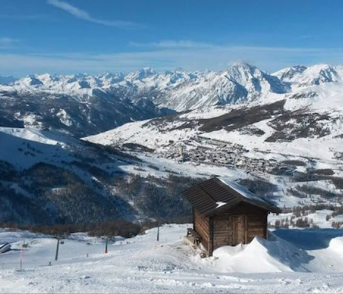 A serene winter scene featuring a snow-covered cabin on a hill, surrounded by ski lifts and mountains under a blue sky.