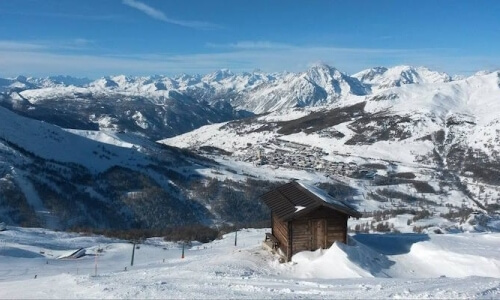 Una cabina di legno immersa nella neve, circondata da alberi e montagne, sotto un cielo blu sereno.