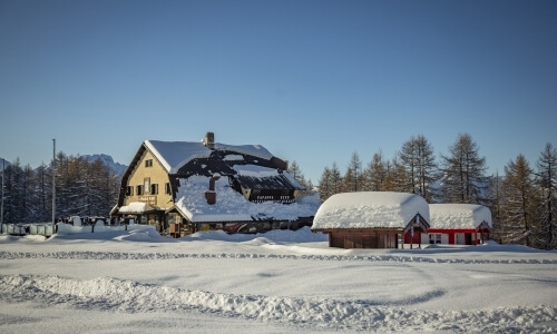 Serene winter scene with a two-story house, red barn, and shed, surrounded by snow-covered landscape and clear blue sky.