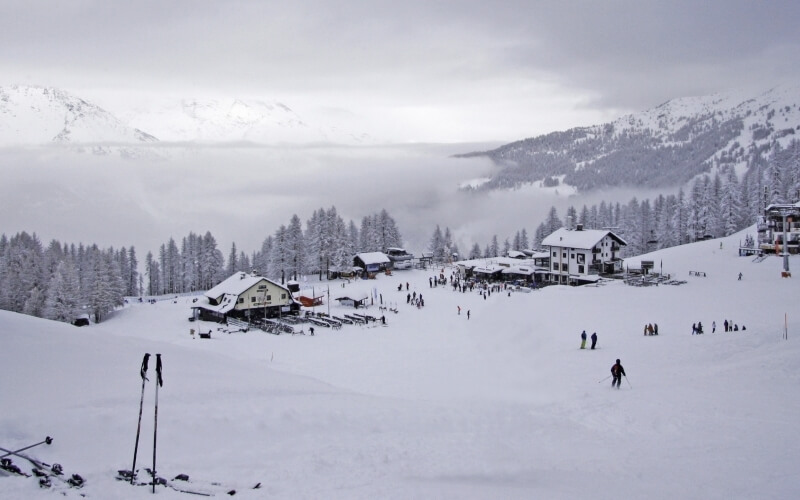 Serene winter scene at a ski resort with snow-covered mountains, trees, buildings, and skiers in the foreground.