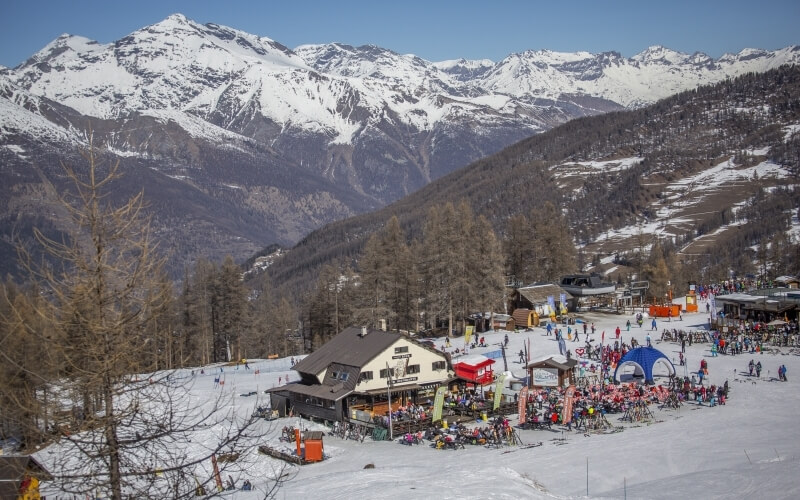 Snowy mountain range with a ski resort, buildings, trees, and people skiing under a clear blue sky.