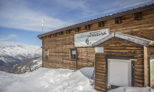 Edificio in legno con tetto inclinato, insegna "osteriafrecura", circondato da neve e montagne, cielo sereno.