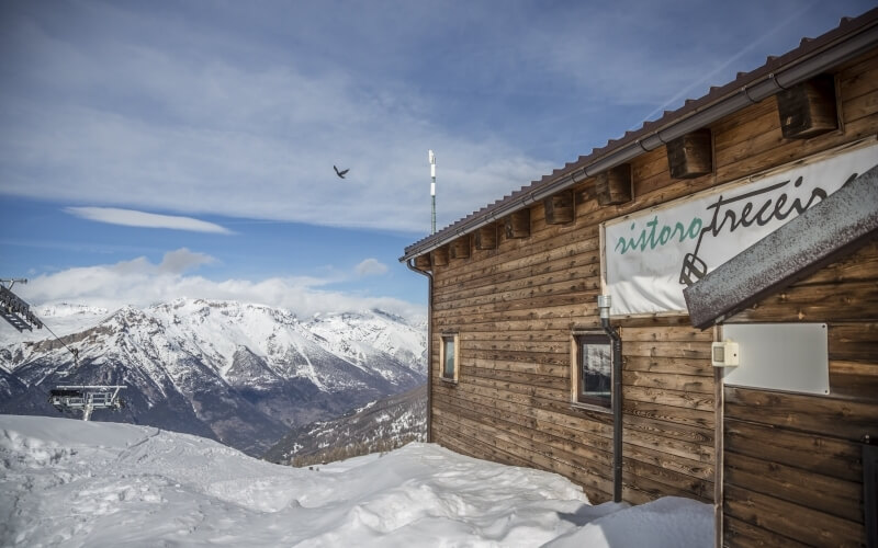 Serene winter scene with a rustic building, snow-capped mountains, ski lift, and a bird in flight against a blue sky.