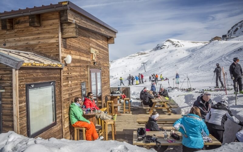 Ski lodge on a snowy mountain with people seated outside and skiers in the background under a blue sky with clouds.