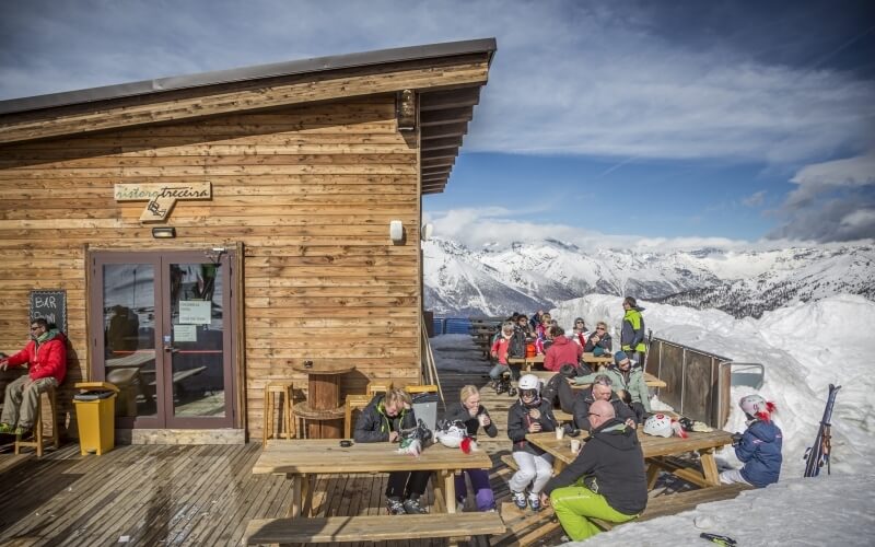 Wooden building "Glory Pretina" on a mountain, with patrons in ski gear and casual attire enjoying food and drinks outside.