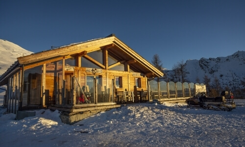 A wooden house with a sloping roof and tractor, set against snow-capped mountains under a clear blue sky.