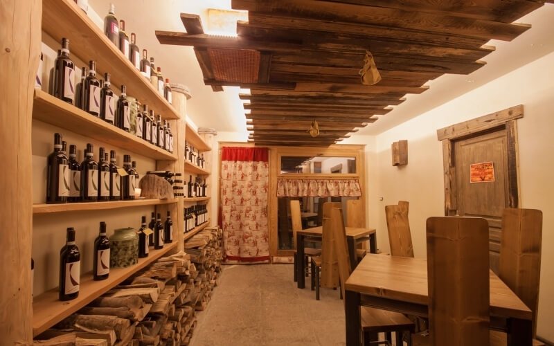 Cozy dining area with wooden shelves of wine, a bar, tables, and chairs, featuring warm wooden accents and inviting decor.