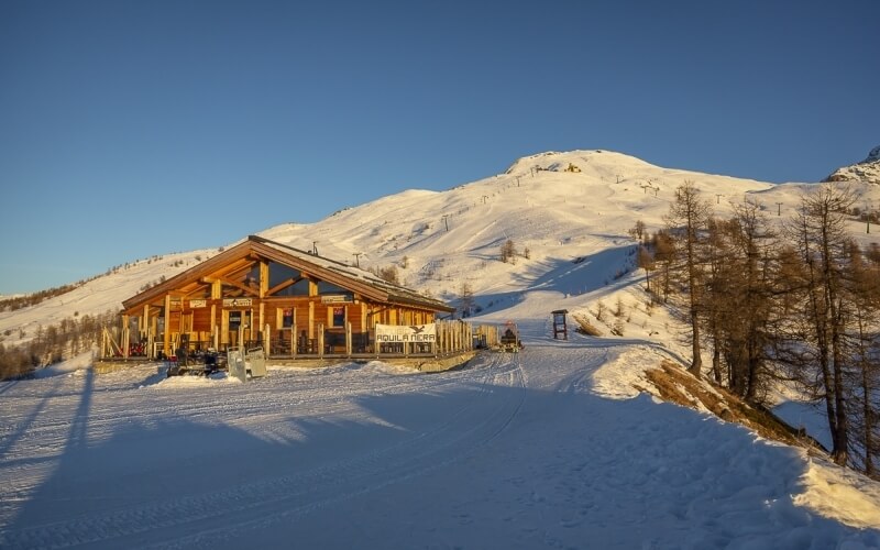 Resort di montagna con edificio A-frame, pista da sci e albe illuminate dal sole, circondato da neve e alberi.