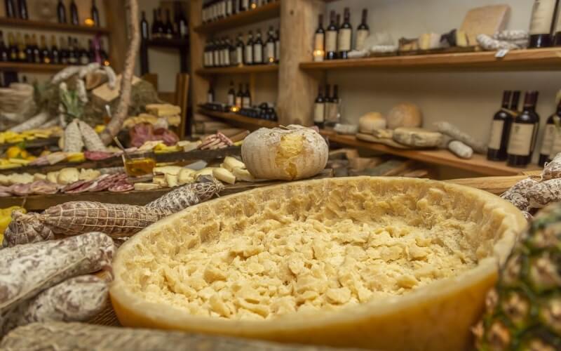 Disposizione attraente di cibo con un grande formaggio al centro, circondato da vini e altri prodotti alimentari.