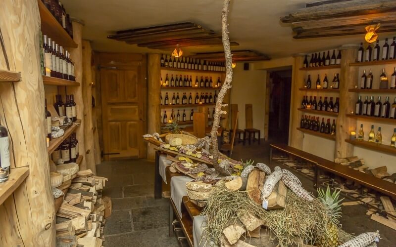 Rustic room with wooden shelves of bottles, a long table of food, and a cozy atmosphere, resembling a winery or tavern.