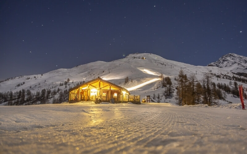 Serene winter scene with a snow-covered mountain, illuminated ski lift, cozy cabin, and starry blue sky.