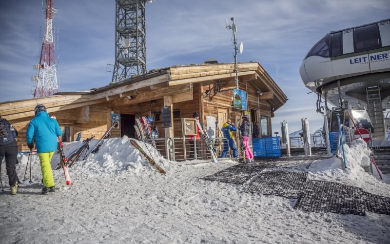 Ski resort scene with a wooden building, people in winter attire, skis, and a chair lift under a blue sky.