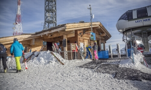 Ski lift station with people outside, snow piles, a LEITNER cable car, and a radio tower against a blue sky.