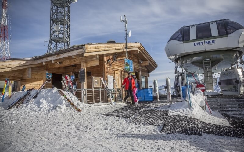 Ski lift station with wooden building, skiers walking, snow-covered ground, and gondola lift in a bustling ski resort.