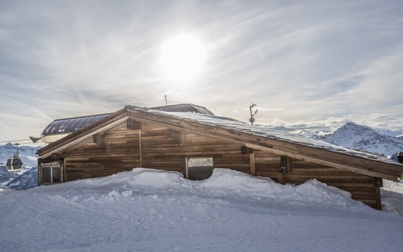 Wooden building on a snowy mountain, gondola lifts nearby, with a scenic view of snow-covered peaks and a bright sky.