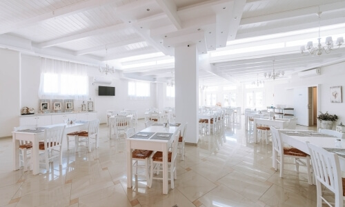 Area pranzo moderna e spaziosa con soffitto a cassettoni, tavoli bianchi, finestre luminose e lampadari eleganti.