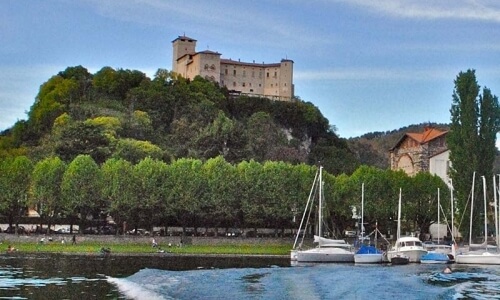 A castle on a hill overlooks calm water with sailboats and a kayaker, surrounded by lush greenery and distant buildings.