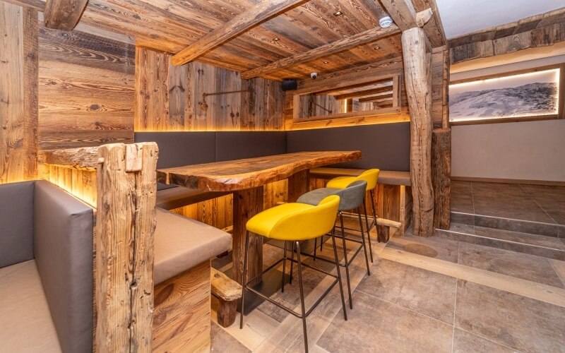 Area di sosta interna in legno e pietra, con sgabelli alti e panchine, illuminata da una luce calda e accogliente.