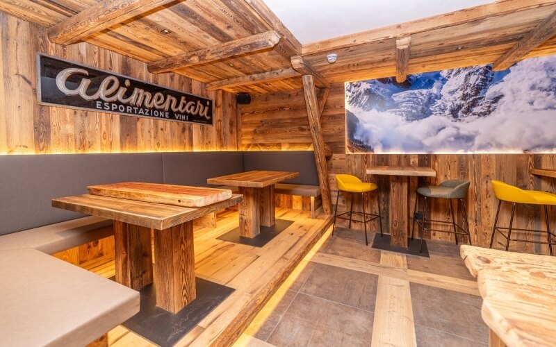 Interior of a rustic restaurant with wooden walls, "Alimentari" sign, snowy mountain poster, and cozy seating arrangements.