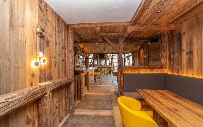 Rustic wood-paneled room with a bar, cozy seating areas, and warm lighting, featuring a central pathway and yellow chairs.