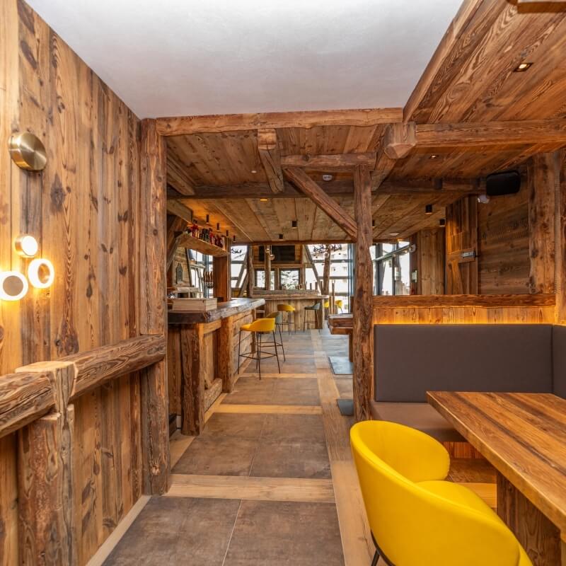 Interno di un ristorante rustico con pareti in legno, corridoio, bar, booth e sedie gialle. Atmosfera accogliente.