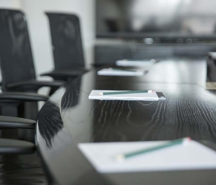 Dark wooden table with chairs, papers, and green pencils in a blurred room with white walls, suggesting a professional setting.