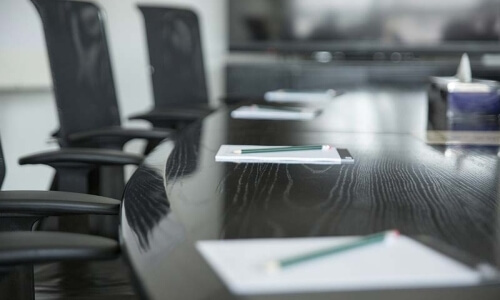 Office chairs around a trapezoid table covered with notepads and pencils, with a distant tissue box on the right.