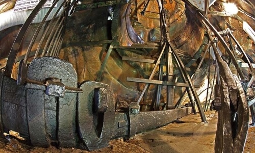 Historical ship with visible damage, wooden structure, metal supports, and a tapestry in the background.