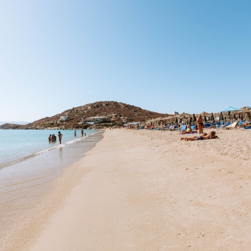 Spiaggia serena con sabbia, acqua calma, cielo blu, persone che nuotano, prendono il sole e si rilassano sotto ombrelloni.