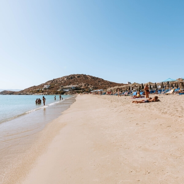 Spiaggia serena con sabbia, acqua calma, ombrelloni blu e bianchi, persone che si rilassano e una collina rocciosa sullo sfondo.