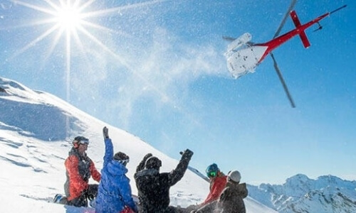 Red helicopter with a white net flies over snowy hills, while six colorful individuals watch against a backdrop of mountains.