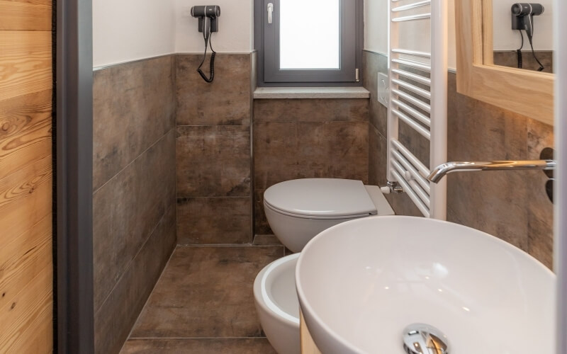 Compact bathroom with brown tiled walls, toilet, sink, hair dryers, and a wooden cabinet with a mirror. Natural light from window.