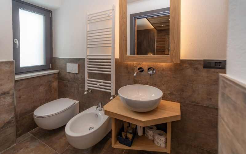 Modern bathroom featuring a toilet, bidet, sink, mirror, and window, with white walls and brown tile flooring.