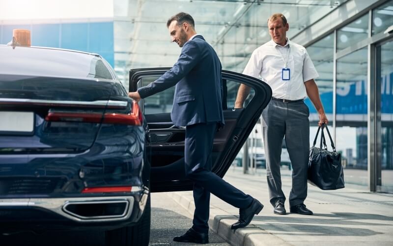 Two men outside a building; one exits a car, the other holds a bag, both dressed professionally, suggesting a business meeting.