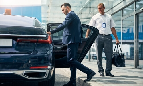 Two men in suits stand by a black car near a modern glass building, one holding a bag and the other by the car door.